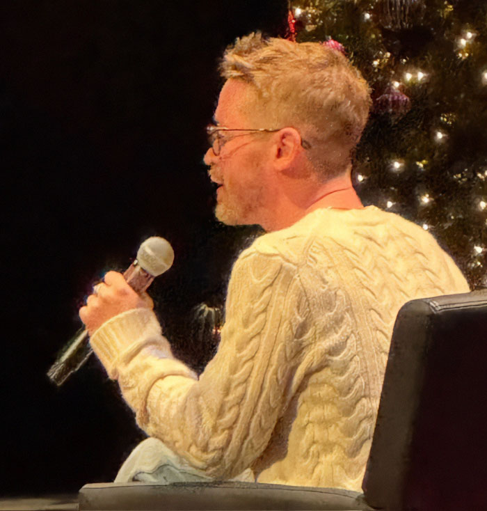 Macaulay Culkin speaking into a microphone while seated, wearing glasses and a cream cable-knit sweater during an event.