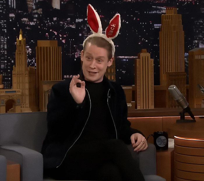Macaulay Culkin wearing bunny ears on a late night talk show, smiling and gesturing with his hand.
