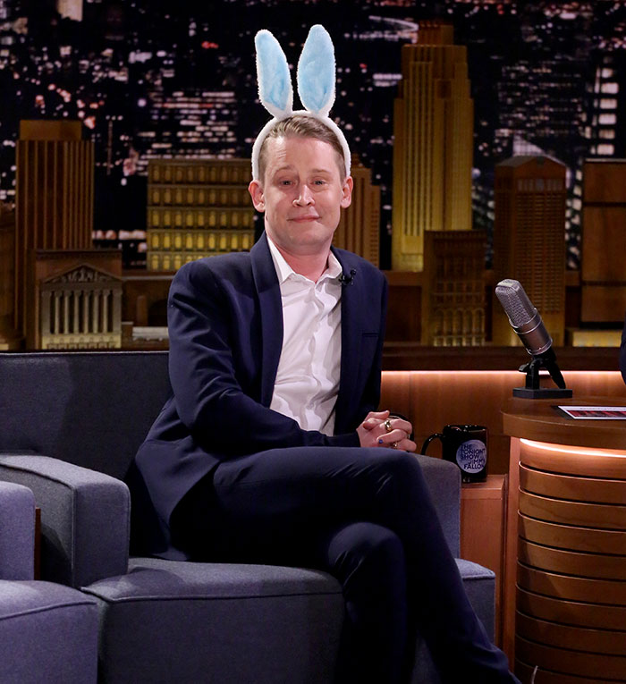 Macaulay Culkin wearing bunny ears and a suit while sitting on a talk show set during an interview.