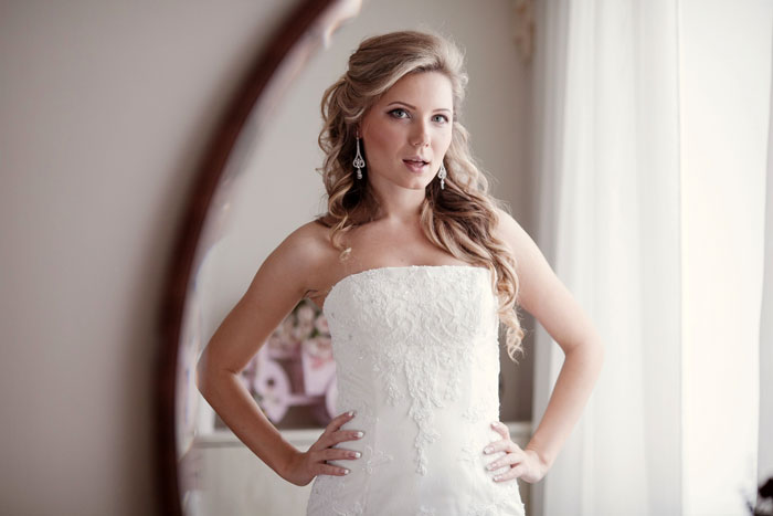 Woman wearing a fancy bridal gown, posing confidently in a well-lit room with soft curtains in the background