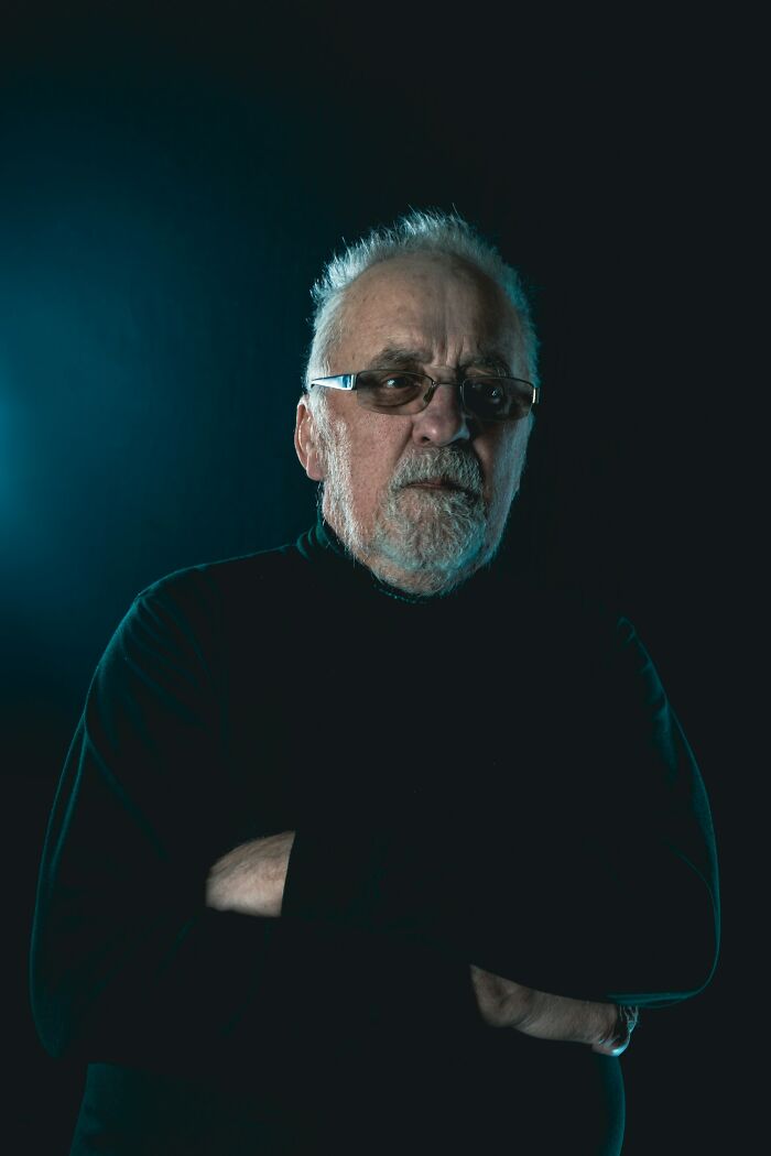 Older man with glasses and beard, arms crossed, in a dark setting conveying heavy confessions and untaken charges.