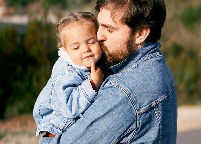 Man in denim jacket holding and kissing young child outdoors, illustrating family struggles with teen's rude attitude.