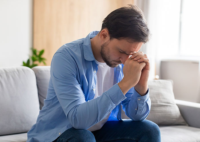 A stressed man sitting on a couch with his head bowed, showing frustration from dealing with a teen&rsquo;s rude attitude.