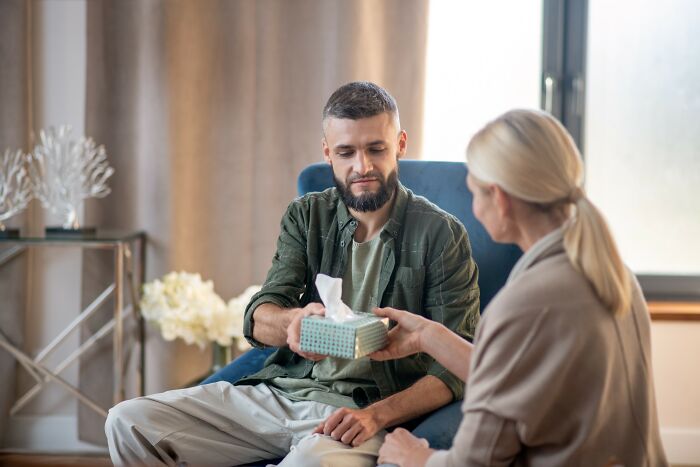 Man holding tissues while woman talks to him in a home setting, illustrating emotional impact of scams on people.