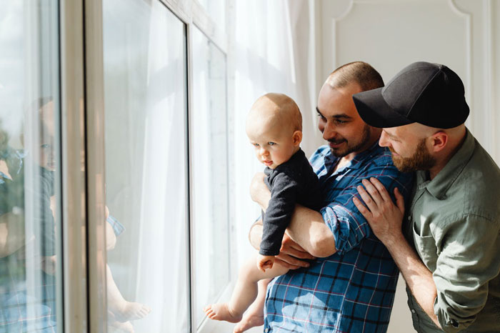 Man holding baby near window with another man standing close by, depicting family surprise on Christmas visit.