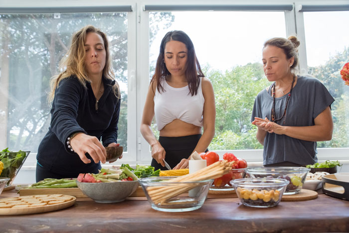 Three women preparing fresh vegetables and snacks together in a bright kitchen setting focused on traditional family roles.