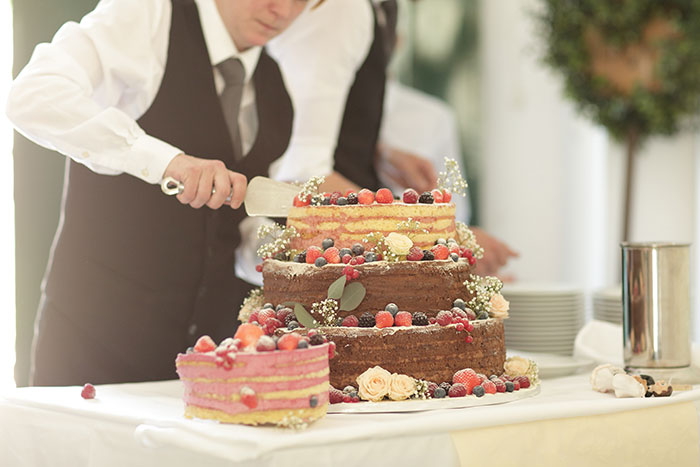 A man in formal attire cutting a multi-tiered cake decorated with berries and flowers at an event.