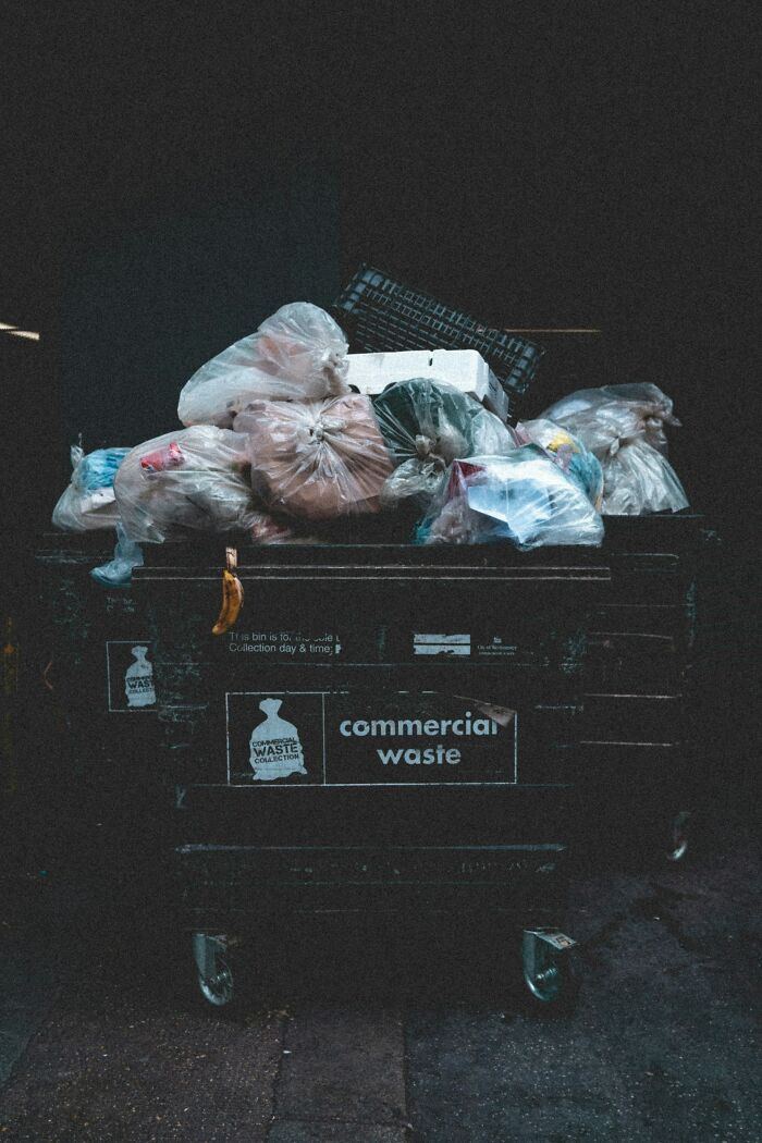 Overflowing commercial waste dumpster filled with tied garbage bags in a dark alley, hinting at garbage men horrors.