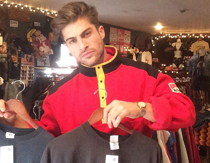 Young man styled like the next Brad Pitt, wearing a red jacket, holding two shirts in a vintage clothing store.