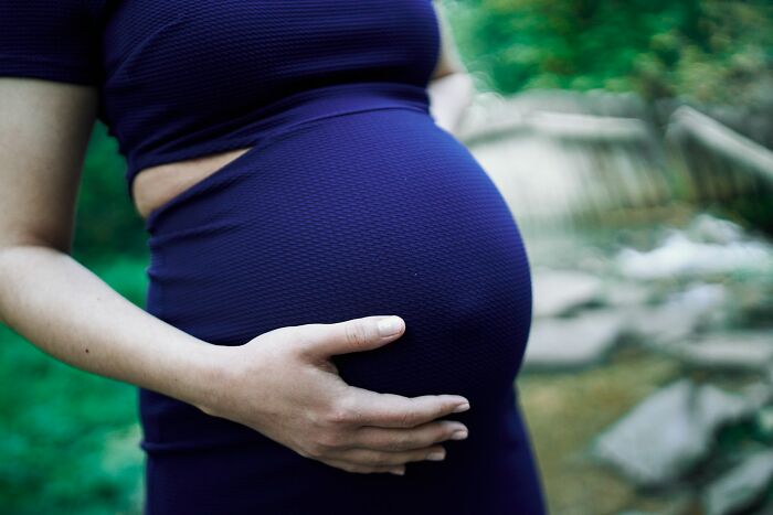 Pregnant woman wearing a dark blue dress, holding her belly in an outdoor setting with blurred nature background.