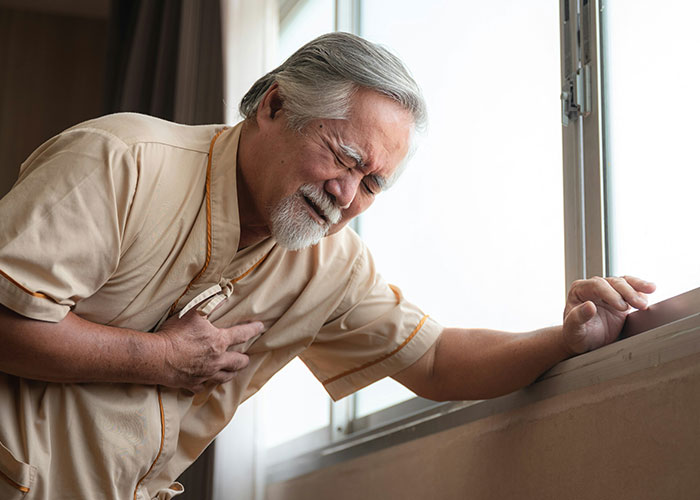 Elderly man clutching chest in pain near window, illustrating dangers in everyday life for medical malpractice lawyer cases.