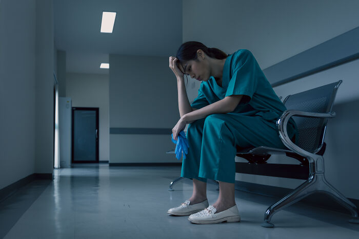 Doctor in scrubs sitting alone in a hospital hallway looking stressed, reflecting on eerie patient encounters.