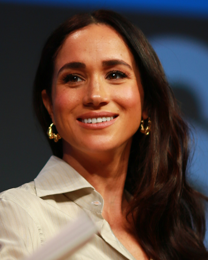Meghan Markle smiling during an event, wearing gold earrings and a cream shirt with an awkward royal gesture discussed. Meghan Markle smiling during an event, wearing gold earrings and a cream shirt with an awkward royal gesture discussed.