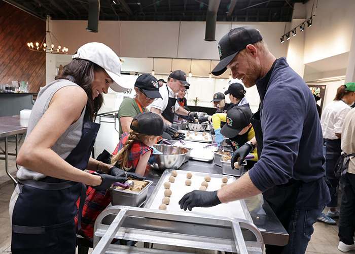 Meghan Markle participating in a volunteer event, involved in an awkward royal gesture with others preparing food. Meghan Markle participating in a volunteer event, involved in an awkward royal gesture with others preparing food.
