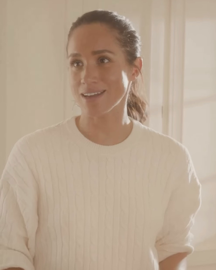 Meghan Markle in a casual white sweater, smiling indoors with warm natural light during a Thanksgiving turkey event.