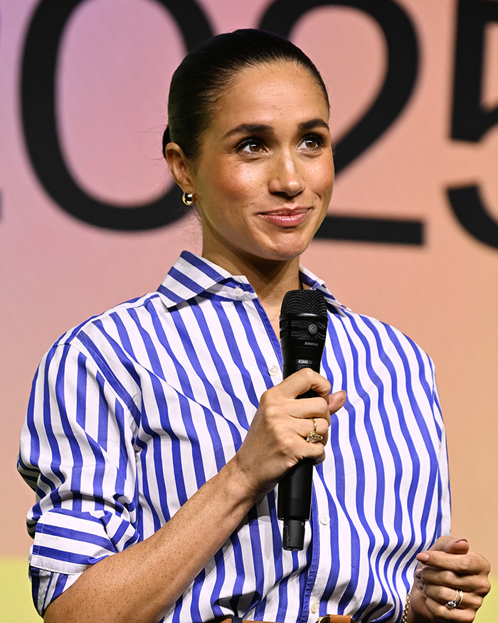 Meghan Markle speaking on stage holding a microphone with a striped blue and white shirt in an event setting.