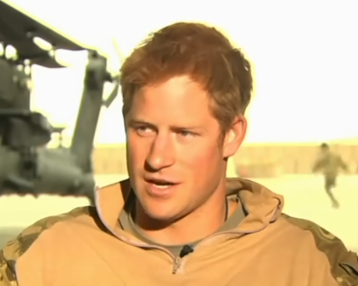 Prince Harry speaking outdoors in military gear with a helicopter and soldiers in the background on Veterans Day.