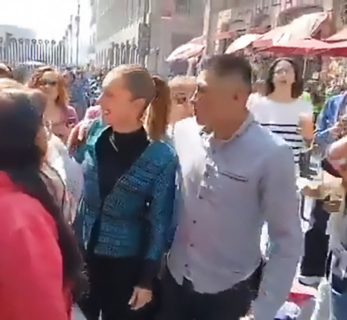 Man kissing and fondling Mexican president while she speaks to crowd with people and buildings in background