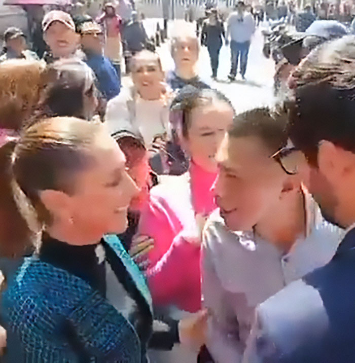 Mexican president speaking to crowd as man kisses and fondles her in public during event.