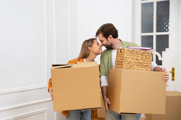 Young couple smiling and holding moving boxes inside a house, depicting family holiday exclusion and conflict. Young couple smiling and holding moving boxes inside a house, depicting family holiday exclusion and conflict.