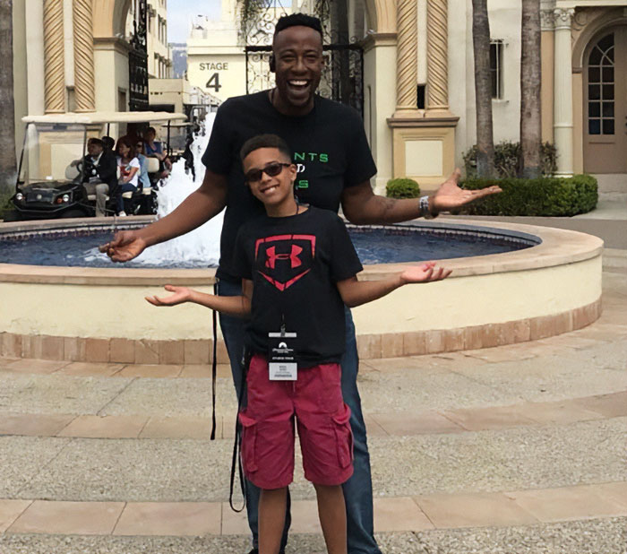 Celebrity chef with dark side standing outdoors with a smiling child near a fountain at a film studio stage.