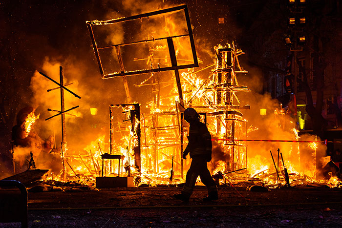 Silhouette of a person walking past a large burning structure at night, capturing a mildly disturbing fact scene.