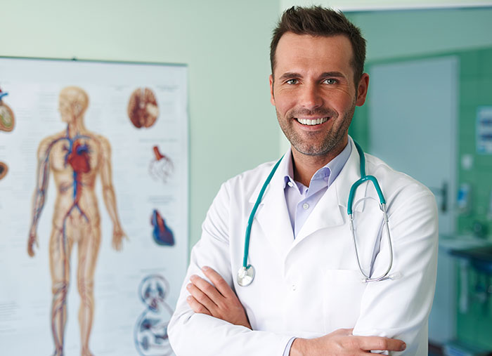 Smiling male doctor with stethoscope standing in medical office near human anatomy poster, representing mildly disturbing fact concept.