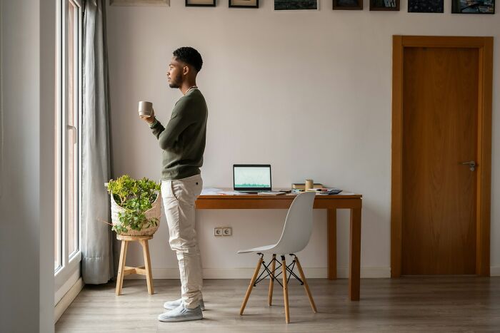 Young man standing near window, holding a cup and reflecting, illustrating subtle signs of cheating in a modern home office.