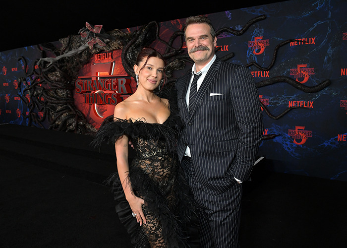 Millie Bobby Brown and David Harbour pose together at Stranger Things Netflix event with dark themed backdrop.