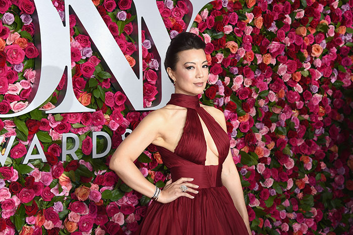 Actress Ming-Na Wen posing in a red gown at an event with a floral wall backdrop highlighting her acting career.