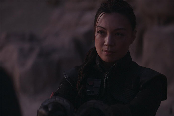 Actress Ming-Na Wen in dark outfit with braided hair, reflecting quietly in a dim, rocky outdoor setting.