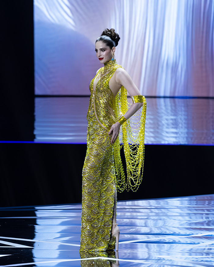 Miss Universe 2025 contestant in a sparkling yellow gown walking on stage during the pageant event.