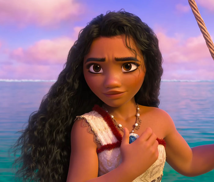 Animated Moana character from Disney's film, with long curly hair and traditional Polynesian attire by the ocean at sunset.