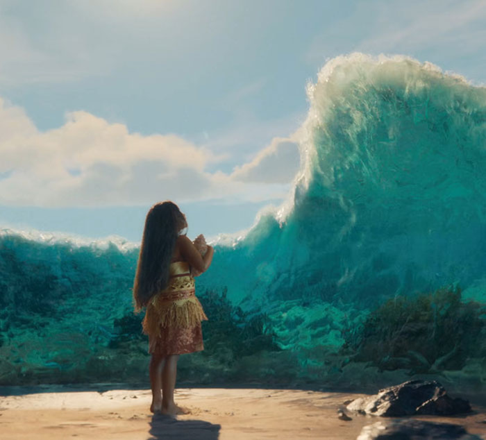 Young girl resembling Moana stands on beach facing large ocean wave in Disney live-action film scene.