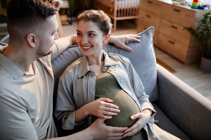 Pregnant woman and man sitting on couch, smiling and holding her belly, related to mom affair college money dad topic.