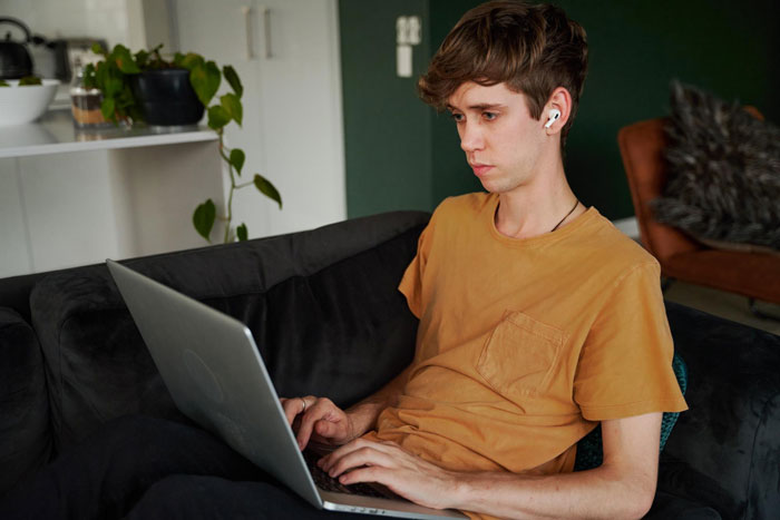 Young man focused on laptop, wearing earbuds, sitting on couch with mom affair college money dad concerns in mind.