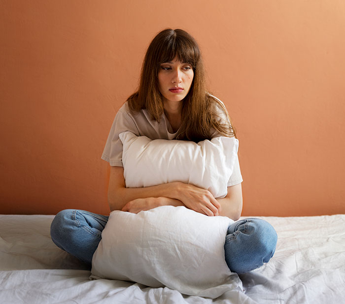Young woman sitting on bed hugging a pillow, reflecting hurt feelings after man cuts ties with mother over texts to girlfriend