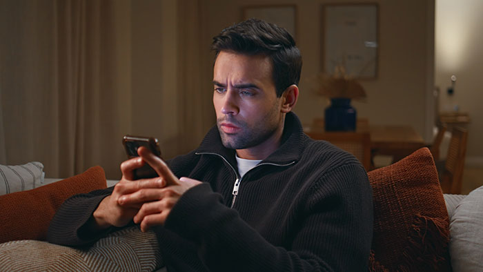 Man sitting on couch in a dark room, looking at his phone with a serious expression, cutting ties with his mother.