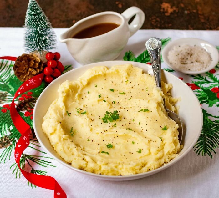 Bowl of mashed potatoes with parsley garnish, gravy boat, and festive holiday decorations on a tablecloth.