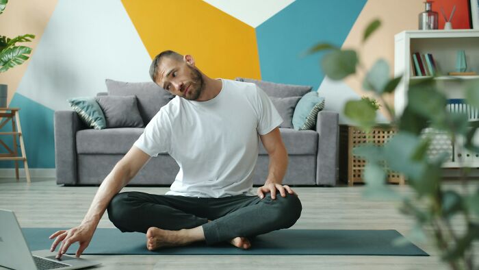 Man sitting on yoga mat stretching neck while using laptop at home, one of the hilarious inventive ways workers got away with breaking rules