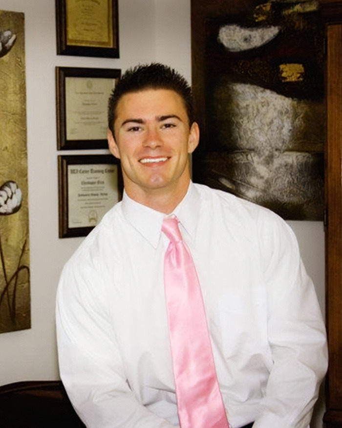 Young man in white shirt and pink tie smiling indoors with framed certificates and artwork in the background about skinned body Las Vegas museum case.