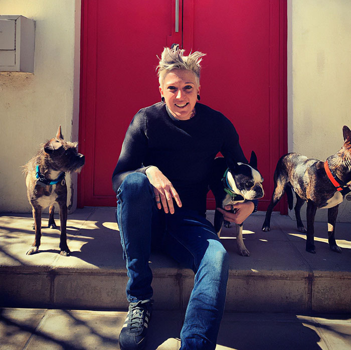 Celebrity chef sitting on steps outside red door, smiling and surrounded by three small dogs on a sunny day.