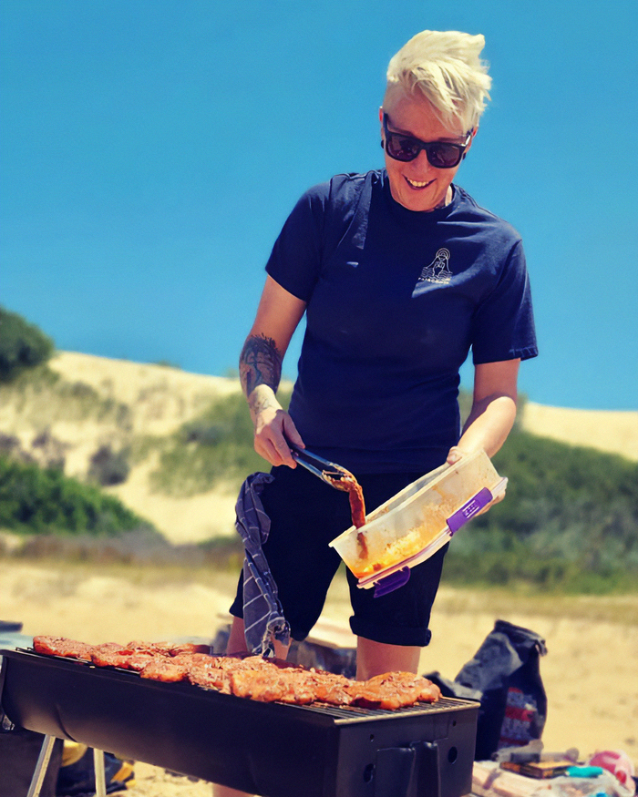 Celebrity chef cooking on a grill outdoors, smiling and preparing food under a clear blue sky in a natural setting.