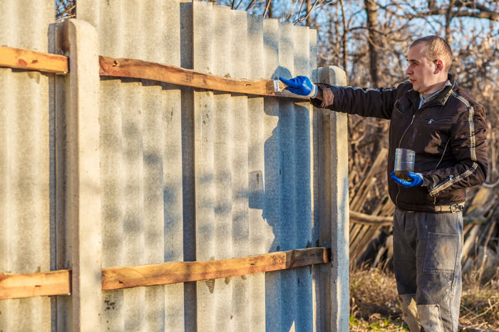 Man inspecting newly replaced fence by neighbor, standing outdoors with paintbrush and wearing blue gloves.