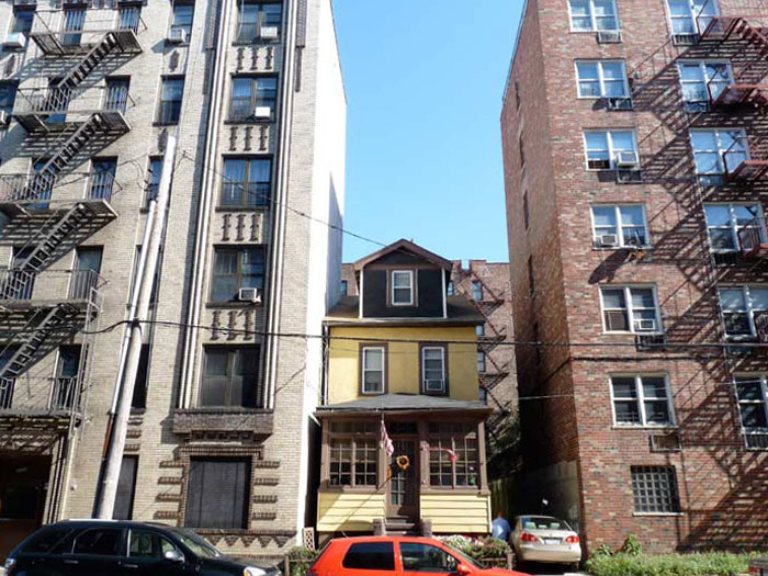 Small yellow house framed by tall concrete and brick apartment buildings with fire escapes and parked cars below. Small yellow house framed by tall concrete and brick apartment buildings with fire escapes and parked cars below.