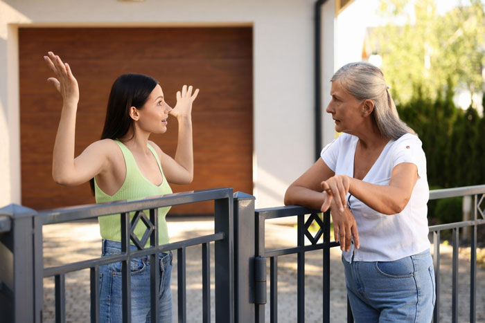 Two women having a tense conversation over a metal fence about a plant wall and a pure concrete view between neighbors. Two women having a tense conversation over a metal fence about a plant wall and a pure concrete view between neighbors.