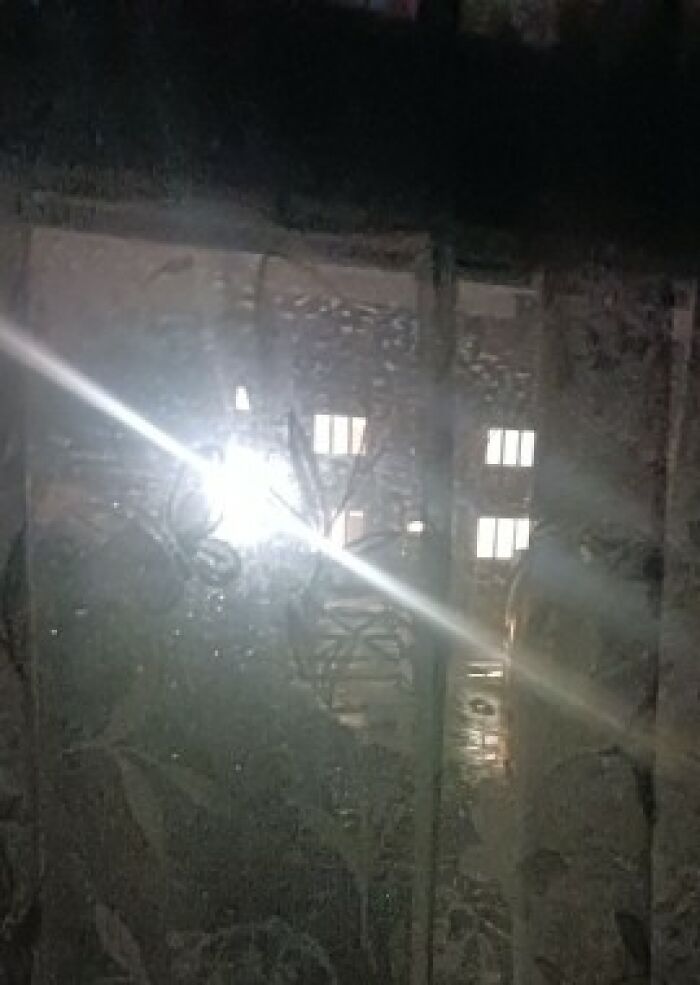 Bright light shining through a patterned curtain at night with illuminated windows from neighbours in the background.