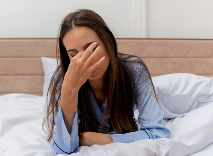 Young woman in blue pajamas on bed covering her face with hand, feeling tired or stressed under actual sun light.