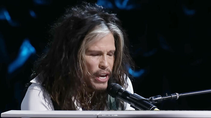 Musician with long hair and white streaks singing passionately into a microphone during a live performance.