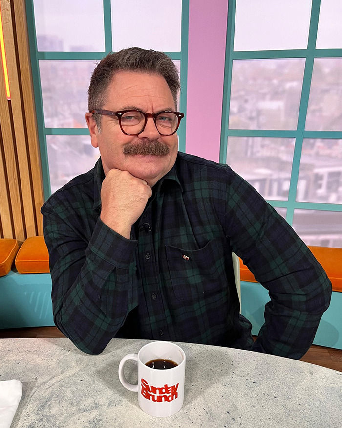 Nick Offerman seated at a table with coffee mug, wearing glasses and plaid shirt, during live talk show broadcast.